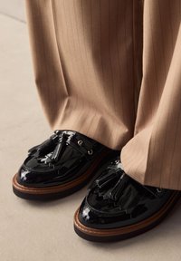 Mocassins en cuir verni noir avec pampilles, bout arrondi et semelle en caoutchouc marron. Associés à un pantalon beige à rayures.