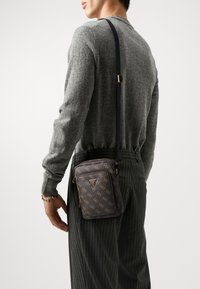 Sac bandoulière marron foncé avec motif logo doré, en cuir, poche zippée, bandoulière réglable et forme rectangulaire. Surface texturée.