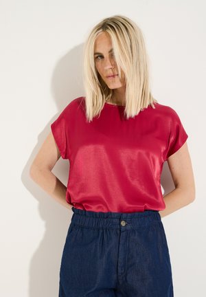 Blonde Frau mit schulterlangem Haar, die ein rotes Satin-Kurzarm-Top und dunkelblaue, hoch taillierte Hosen vor einfarbigem Hintergrund trägt.