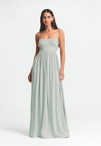Šviesiai žalia maxi suknelė, turinti plonas petnešėles, prigludusią liemenį su raukiniais ir laisvai krintančią, susiraukšlėjusią sijoną, suteikiančią minkštą, tekstūrinę išvaizdą.