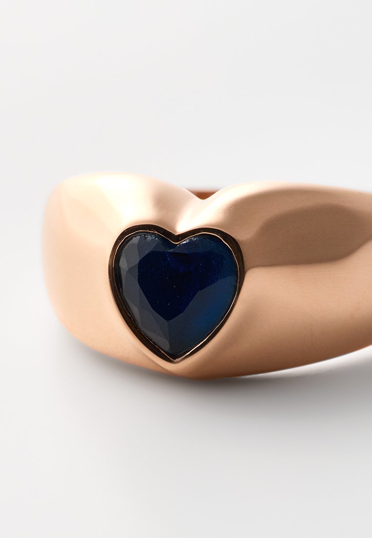 Anillo de oro rosa con una gema azul en forma de corazón facetada, enmarcada por un contorno negro. Banda lisa y redondeada, con acabado pulido.