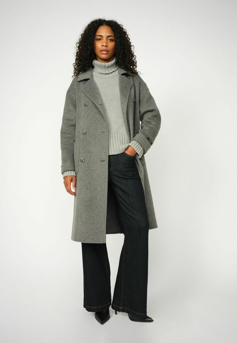 Cappotto grigio in misto lana con bottoni doppi, ampi rever, abbinato a un dolcevita a costine grigio chiaro e jeans scuri a gamba larga.