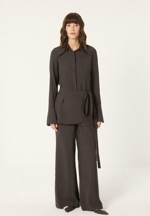 Femme aux cheveux mi-longs portant un haut à manches longues gris foncé avec de larges poignets, un pantalon large assorti et un détail noué sur le côté.