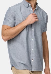 Camisa de botón de manga corta en tela azul claro, con un emblema bordado, botones blancos y un corte ajustado con aberturas laterales.