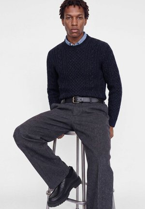 Un mannequin masculin assis porte un pull bleu marine à torsades par-dessus une chemise bleue, un pantalon gris, une ceinture noire et des chaussures noires, devant un fond de studio uni.