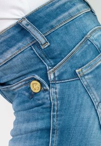 Jeans en denim bleu clair ; dotés d'un bouton doré avec un logo, de surpiqûres contrastées et d'une texture lisse avec de légères zones de distresse.