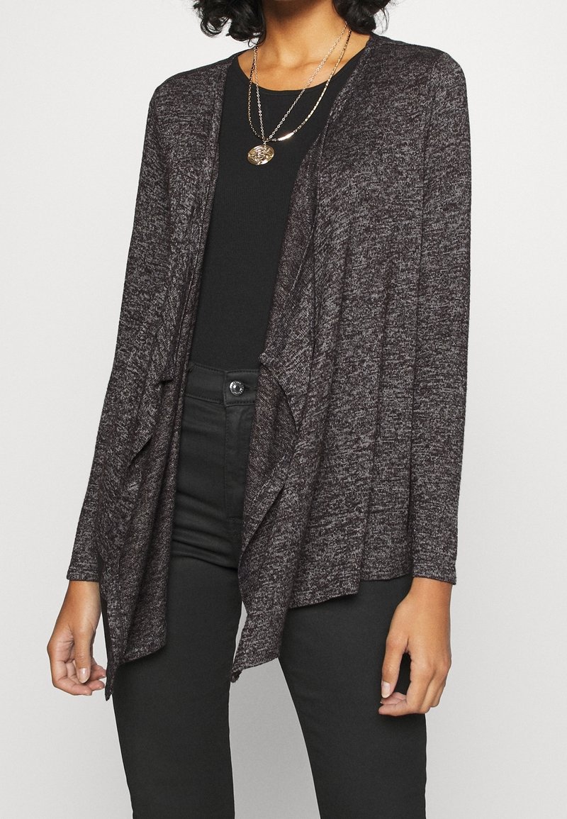 Tmavě šedý kardigan z texturované pleteniny, s otevřeným předním dílem, dlouhými rukávy a volným střihem. Nošený přes černé tričko.