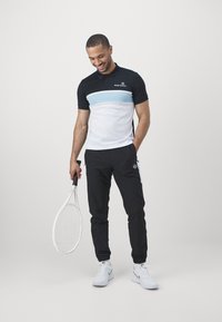Pánsky športový outfit pozostávajúci z čierno-bielej poloskie, s svetlomodrým pruhom, v kombinácii s čiernymi nohavicami a bielymi teniskami, drží raketu.