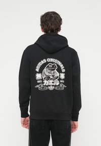 Sweat à capuche noir en coton avec une poche kangourou à l'avant. Présente un grand motif blanc de grenouille au dos, avec des accents de texte et un logo.