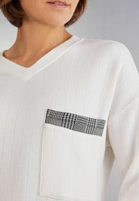Top blanco de cuello en V con textura de canalé vertical y un detalle decorativo de bolsillo a cuadros en blanco y negro. Telas suaves con un corte relajado.