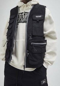 Czarna praktyczna kamizelka z wieloma przednimi kieszeniami, siateczkowymi ramionami i zamkami błyskawicznymi, noszona na szarej bluzie z kapturem i czarnych spodniach.