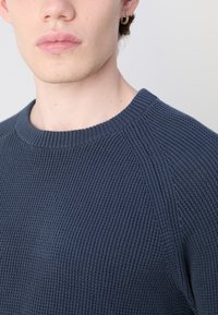 Maglione a maglia blu navy con texture a coste, collo rotondo e maniche raglan, che mette in mostra una vista ravvicinata del collo e della cucitura della spalla.