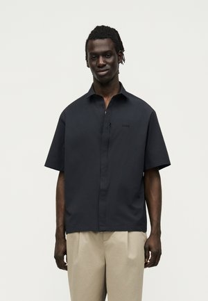 Νεαρός άνδρας με σκουρόχρωμο δέρμα και dreadlocks, φορώντας ένα άνετο μαύρο πουκάμισο με κοντά μανίκια και μπεζ παντελόνι, στέκεται μπροστά σε απλό φόντο.