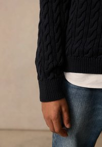 Mornarsko modri pulover z pleteno strukturo in rebrastimi manšetami, v kombinaciji z belo majico in jeansom. Poudarek na teksturi in zapletenih detajlih tkanja.