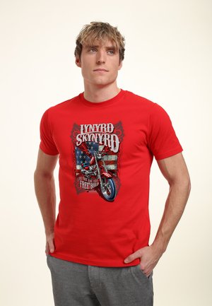 Jonge man draagt een rood Lynyrd Skynyrd T-shirt met een motorfiets- en Amerikaanse vlag ontwerp, staand met handen in de zakken van een grijze broek.