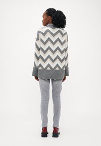 Maglione a zigzag grigio e crema con maniche lunghe, scollo rotondo e orlo a coste, abbinato a pantaloni grigio chiaro e tacchi neri.