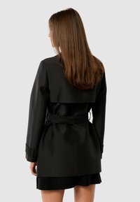 Woman with long brown hair wearing a black belted trench coat facing away against a plain light background.