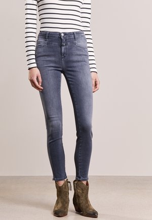 Kvinde iført højtaljede, falmede blå skinny jeans, stribet langærmet top og olivengrønne ankelstøvler, der står på en lys gulv.