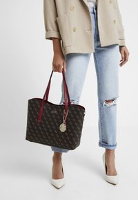 Zwarte totebag met bruin logo-patroon, rode handvatten en zilveren bedel. Draagt lichtblauwe gescheurde jeans en beige oversized blazer.