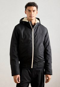 Giacca nera imbottita con zip frontale, cappuccio e fodera crema. Presenta polsini elasticizzati e un orlo aderente, che mette in risalto un design pulito e un accento con logo.
