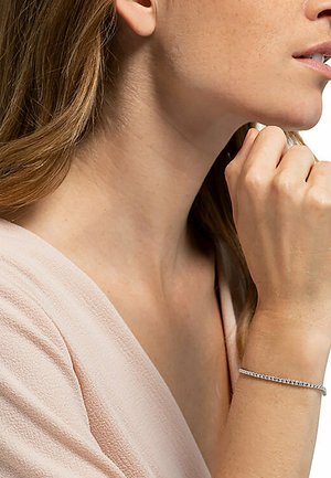Silberarmband mit einem zarten, symmetrischen Design, das kleine runde Steine aufweist. Am Handgelenk getragen, akzentuiert von einem weichen, strukturierten Stoffhintergrund.