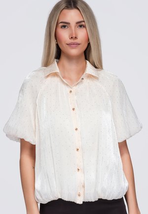 Femme aux cheveux blonds raides porte un chemisier blanc transparent avec des manches bouffantes et de petits pois, debout devant un fond clair uni.