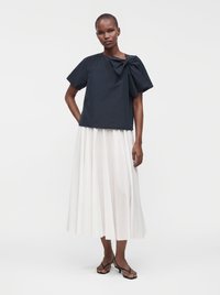 Femme debout portant un haut noir à manches courtes avec un grand nœud sur l'épaule, une jupe midi plissée blanche et des sandales noires à talons bas.