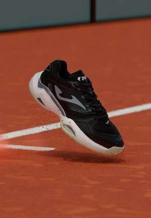 Scarpa sportiva bianca e nera sospesa sopra un campo da tennis in terra rossa con piccole particelle di argilla che volano vicino alle linee bianche del campo.