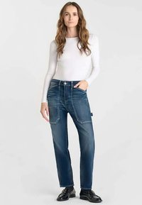 Blauwe spijkerbroek met een relaxte pasvorm, voorzien van grote opgestikte zakken en contrasterende stiksels. Gecombineerd met een wit shirt met lange mouwen.