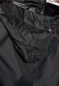 Veste à capuche noire avec des coutures visibles et un tissu légèrement froissé, montrant un détail élastique sur le bord de la capuche.