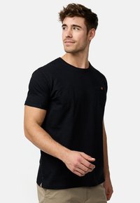 Schwarzes Baumwoll-T-Shirt mit Rundhalsausschnitt, kurzen Ärmeln und einer kleinen Brusttasche mit Knopfdetails. Seitenschlitze am Saum.
