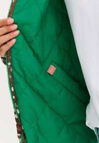 Wyściółka pikowanej zielonej kurtki o gładkiej teksturze, z różową metką oraz brązowym wykończeniem z wzorzystym akcentem wzdłuż krawędzi.
