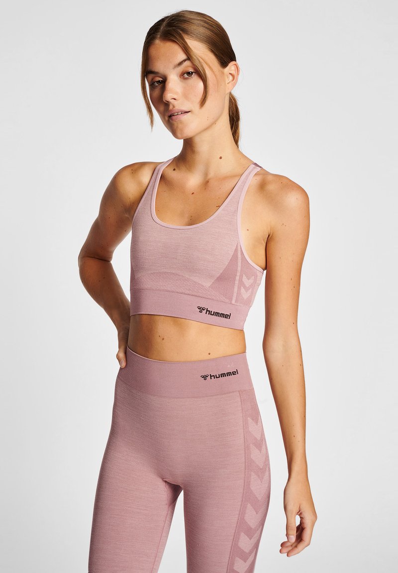 Ljust rosa sportbh och leggings-set, med en texturerad yta, och Hummel-logotyp. Designen inkluderar en subtil chevronmönster längs sidorna.