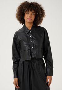 Melnā īsā jaka no mākslīgās ādas, ar apkakli, pogu aizdari, divām priekšējām kabatām un garām piedurknēm ar elastīgām aprocēm.