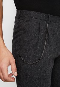 Grijze broeken met een houndstooth-patroon en een gestructureerde afwerking, met een decoratieve zilveren ketting aan de zak.