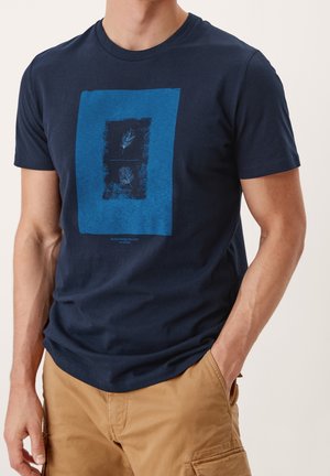 Mand iført en marineblå t-shirt med en blå rektangel og to fjer illustrationer, kombineret med beige cargoshorts, hånd i lommen.