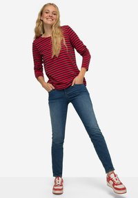 Punainen ja laivastonsininen raidallinen pitkähihainen paita yhdistettynä sinisiin skinny-farkkuihin ja punaisiin lenkkareihin, joissa on valkoisia yksityiskohtia. Malli seisoo rentona.