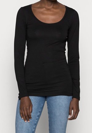 Woman wearing a fitted black long-sleeve scoop neck top and light blue denim jeans, standing against a plain white background.