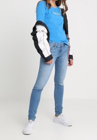 Camisa de manga larga azul con logotipo, chaqueta bomber en blanco y negro, jeans ajustados de mezclilla clara y zapatillas blancas. Conjunto casual con un diseño moderno.