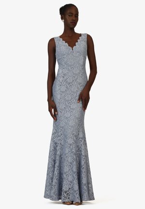 Frau in ärmellosem, bodenlangem, figurbetontem hellblauem Spitzenkleid mit gezacktem V-Ausschnitt, stehend vor weißem Hintergrund.