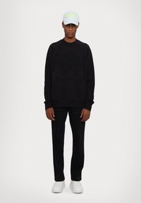 Sort sweater med præget logo, sorte bukser, hvide sneakers og en lyseblå kasket med ordet "VERSACE" i neongul.