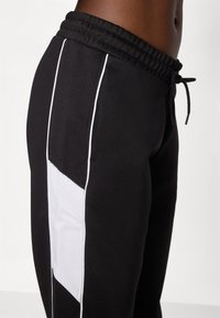 Calças de fato de treino pretas com um painel lateral branco, cintura elástica e cordão. Tecido suave com uma textura lisa. Corte e design padrão.