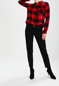 Persona che indossa una camicia a scacchi rossi e neri, pantaloni neri attillati e stivali neri con tacco alla caviglia, con una mano che tocca il viso.