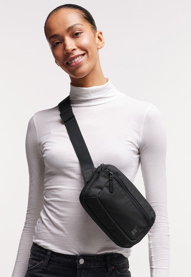 Mujer con un jersey de cuello alto blanco y bolso negro cruzado, sonriendo con el cabello recogido hacia atrás contra un fondo claro y uniforme.