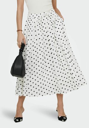 Femme portant une jupe midi blanche à pois noirs, tenant un sac à main noir, assortie à des escarpins à pois noirs et blancs.
