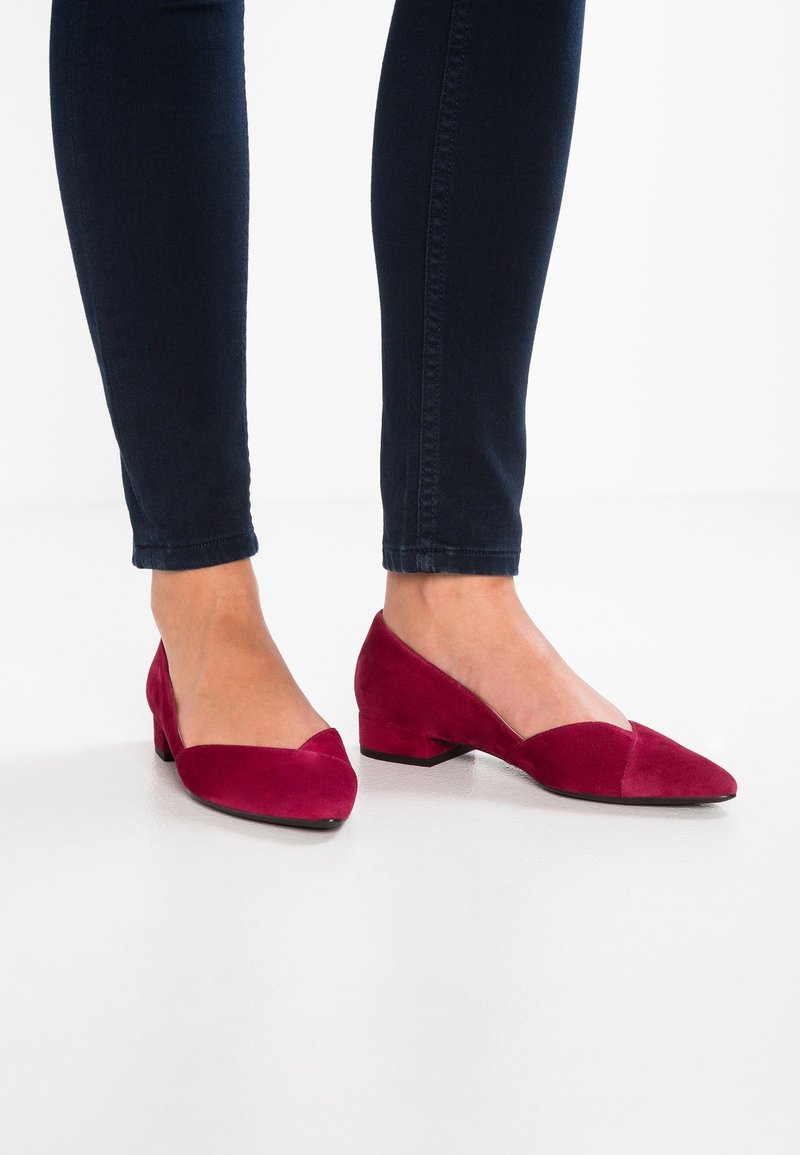 Pieds portant des chaussures à petit talon carré en daim bordeaux, associées à un jean skinny noir sur un fond de sol blanc.