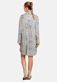 Vrouw met gevlochten haar, gekleed in een lange mouwen jurk tot de knie met een kleurrijk paisley patroon, beige sokken en bruine sloffen.