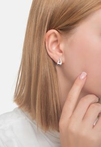 Silverfärgad droppformad studörhänge med en slät yta och en kontrasterande inre accent. Bärs av en modell som framhäver designen mot hennes hår.