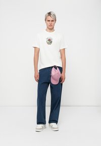 White graphic t-shirt with a colorful print, loose blue denim jeans, and a pink cap resting on the side. White and beige sneakers.