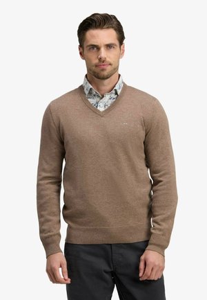 Homme portant un pull col en V marron clair par-dessus une chemise blanche à fleurs avec un pantalon noir, debout devant un fond uni.